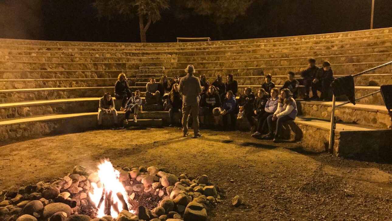 teens listening to a man while sitting around a camp fire.